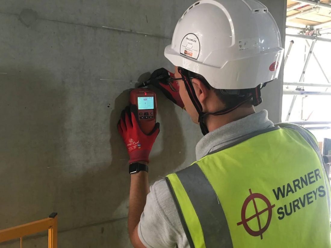 A surveyor holding a cover meter against a concrete wall, reading the rebar behind the surface