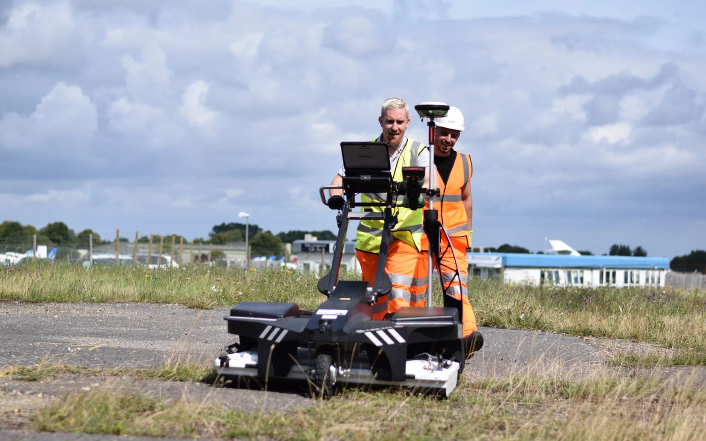 Two surveyors pushing a ground-penetrating radar array across open ground beside a service road
